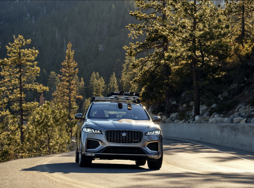 A silver Jaguar SUV driving on a snowy mountain road. The vehicle has a roof rack and is splashing snow as it moves